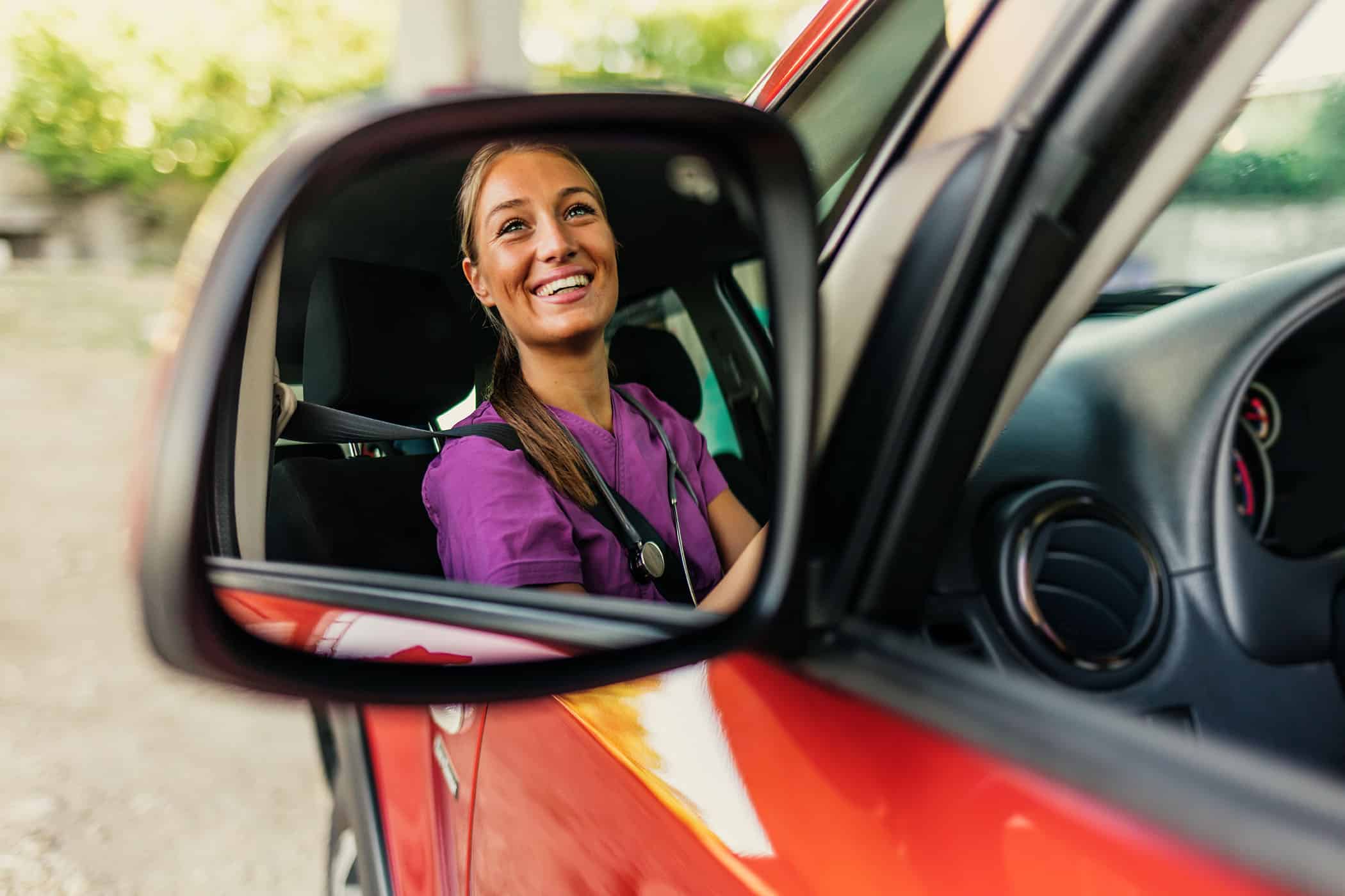 Close up of a reflection in rear-view mirror of young nurse driving a car, smiling.